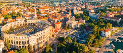 An aerial view of a city with a prominent historical amphitheater, a mix of modern and traditional architecture, and a marina with boats.