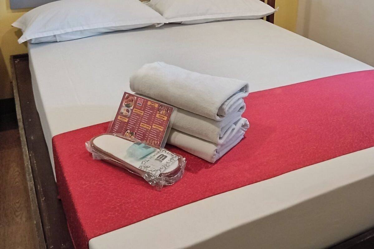 Uma cama com colcha vermelha, duas almofadas, cabeceira de madeira e uma pequena mesa com menu e toalhas.