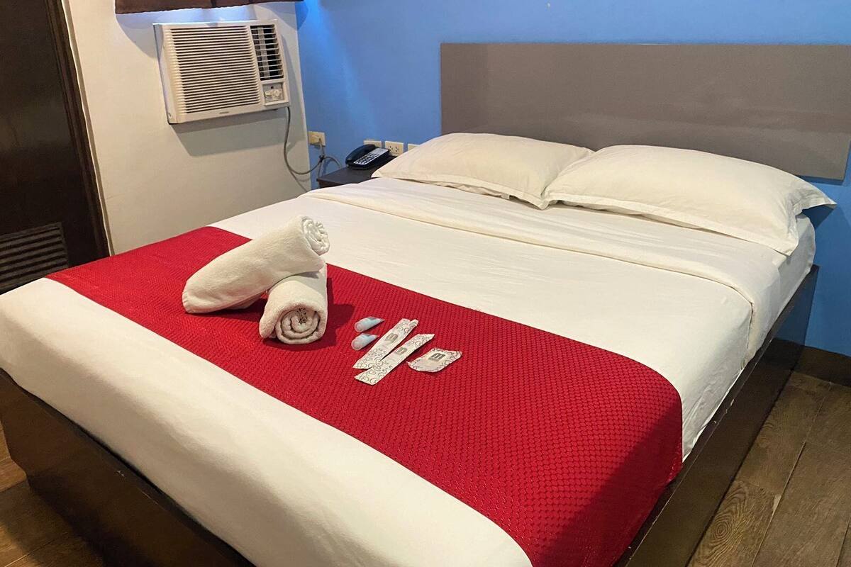 Quarto de hotel com cama, manta vermelha, travesseiro branco e ar condicionado fixado na parede.