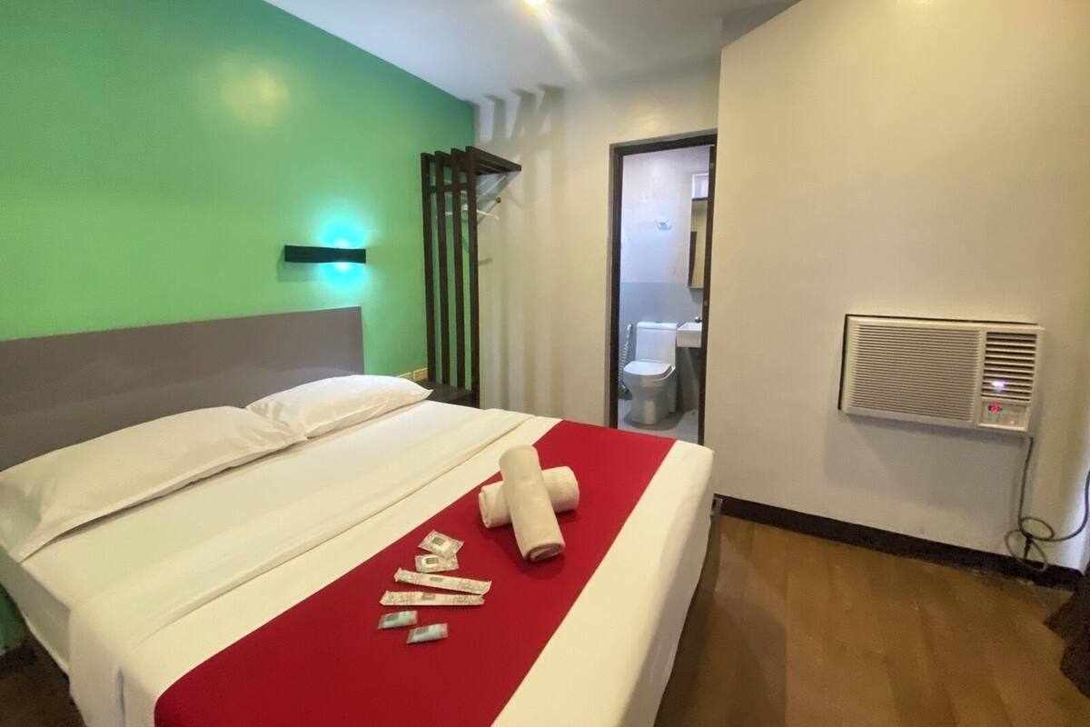 Quarto de hotel com uma cama, uma parede verde, teto branco e banheiro visível por uma porta aberta.