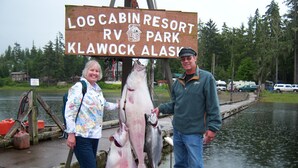 A wooden signboard with text indicating a location, two people standing next to a dock with fish, and a body of water in the background.