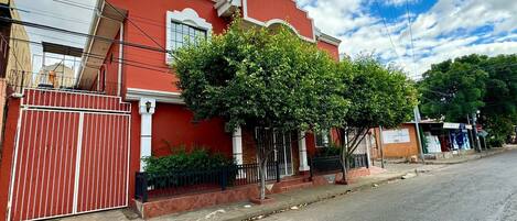 Un edificio rojo con una entrada protegida por una reja, un árbol frente a él y un cielo despejado.