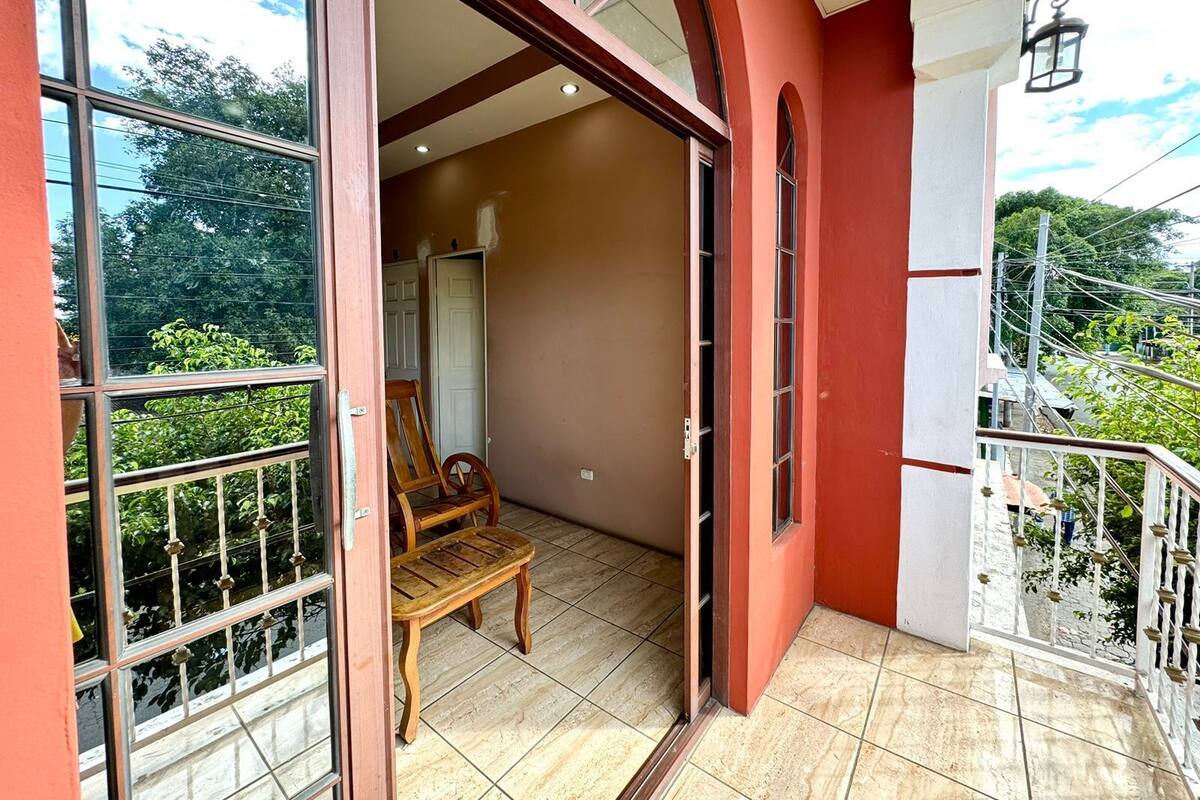 A balcony with a wooden chair, a door leading to an interior room, and a view of greenery.