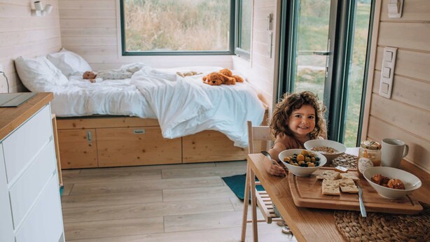 L’intérieur chaleureux d’un chalet avec un lit, un coin repas où se trouve de la nourriture et une fenêtre donnant sur un paysage agréable.