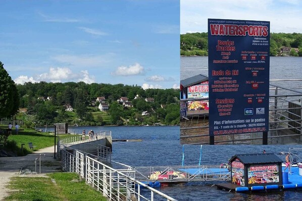 Een watersportfaciliteit met een bord waarop activiteiten en prijzen staan, een steiger met een klein gebouwtje en een meer met huizen op de achtergrond.