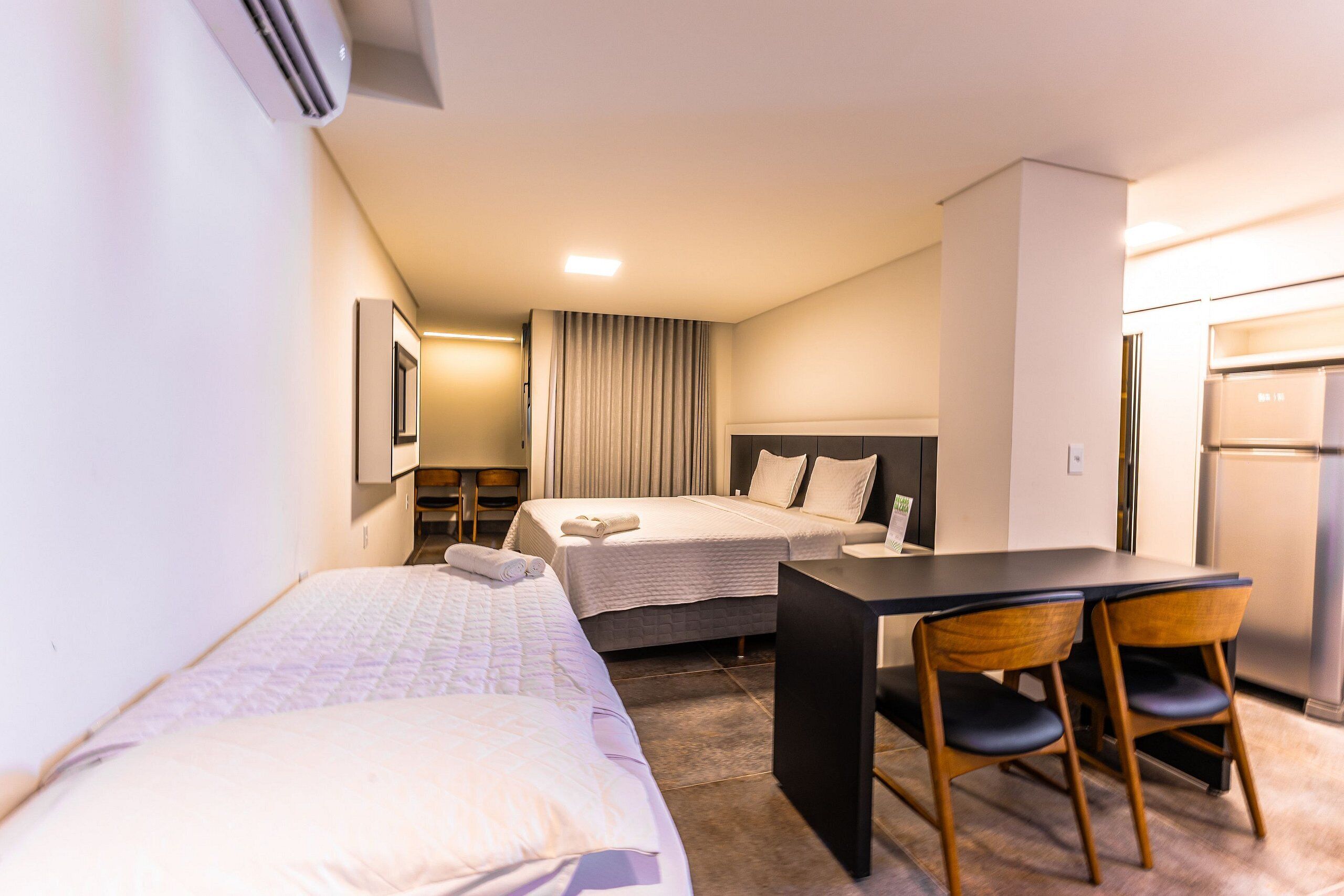 A hotel room with two beds, a desk, chairs, a refrigerator, and a ceiling-mounted air conditioner.