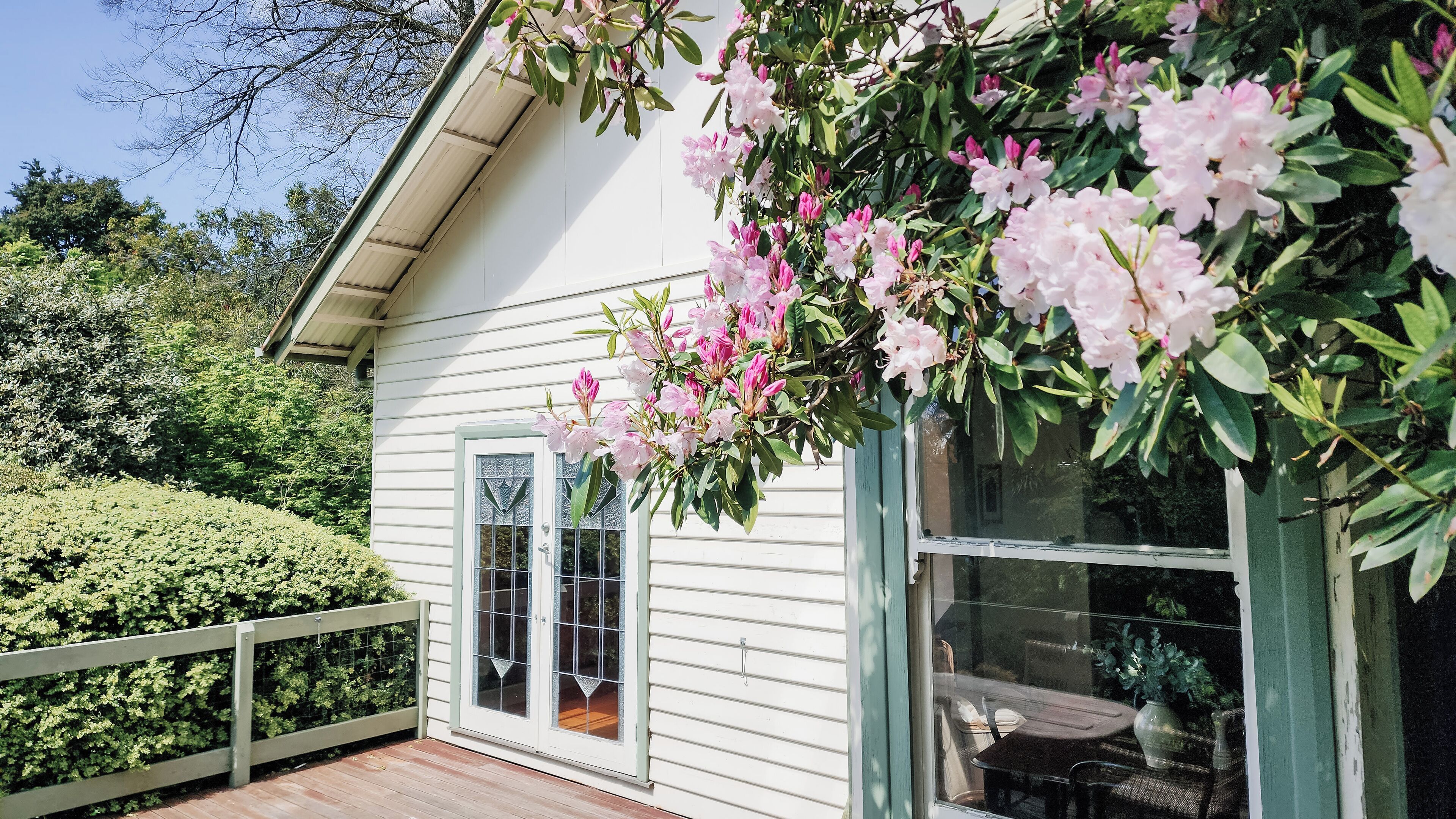 A white house with a wooden deck, a glass door, and pink flowers.