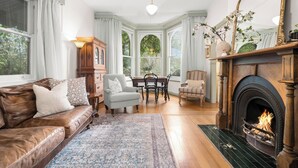 A cozy living room with a leather sofa, a fireplace, a dining table with chairs, and a bay window with curtains. - Woolrich Cottage at Woolrich Estate (Olinda)