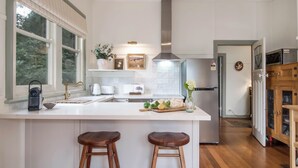 A modern kitchen with white cabinetry, a large island with stools, a stainless steel refrigerator, and a view of the outdoors through large windows. - Woolrich Cottage at Woolrich Estate (Olinda)