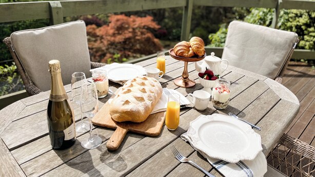 A wooden deck with a table set for breakfast, including croissants, strawberries, and a bottle of champagne. - Woolrich Cottage at Woolrich Estate (Olinda)
