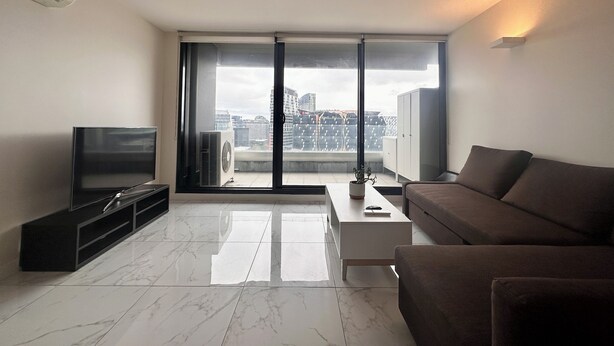 A modern living room with a flat-screen TV, a sofa, a coffee table, and a sliding glass door leading to a balcony with a city view.