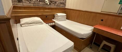 Una habitación de hotel pequeña con cabecera de madera, cama, escritorio y un taburete. En la pared hay cuadros enmarcados y un estante con toallas.