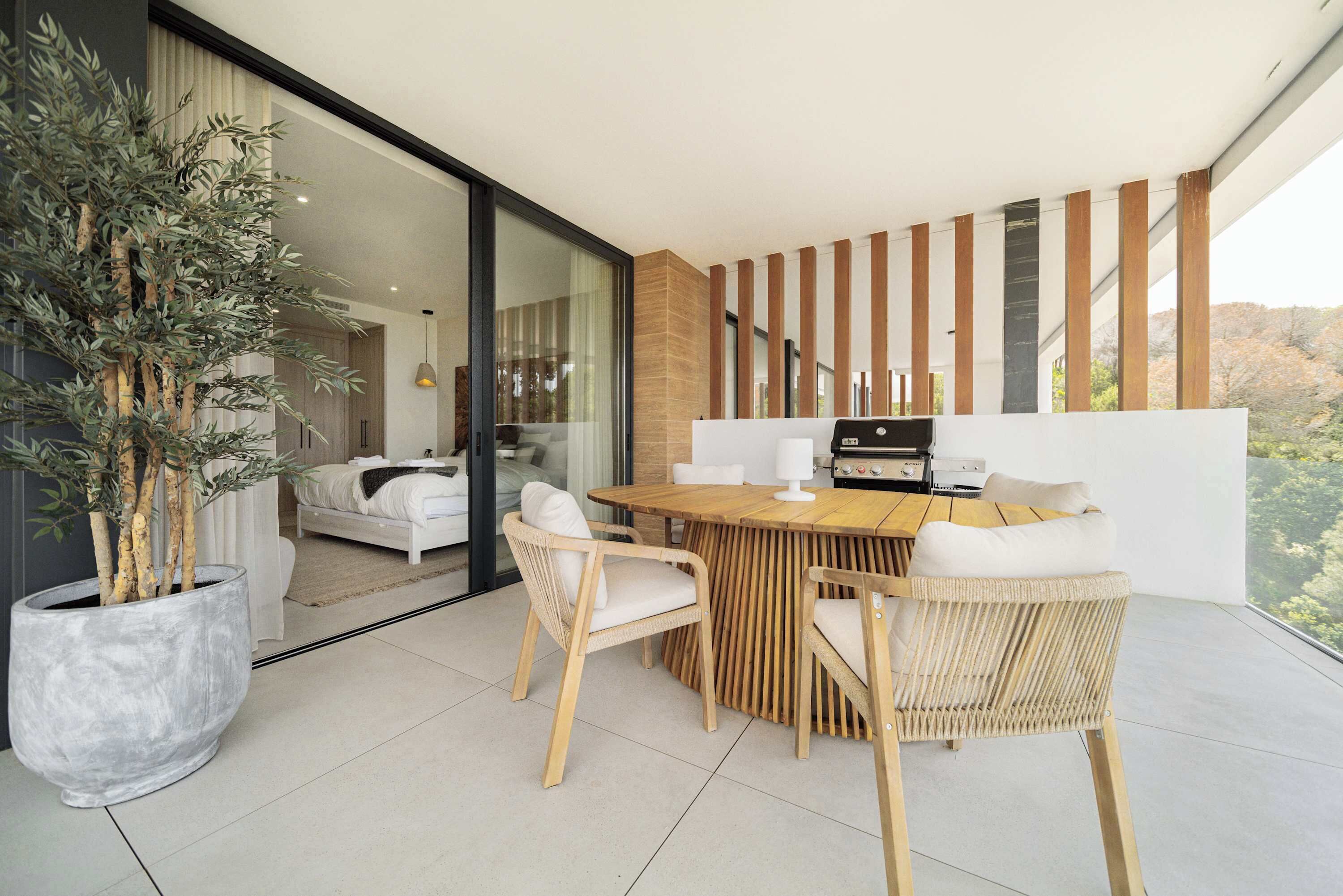 Un espacio moderno para comer al aire libre con una mesa de madera, sillas de mimbre, una planta grande en maceta y vistas a un dormitorio a través de puertas de cristal.