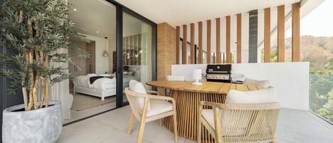 Un espacio moderno para comer al aire libre con una mesa de madera, sillas de mimbre, una planta grande en maceta y vistas a un dormitorio a través de puertas de cristal.
