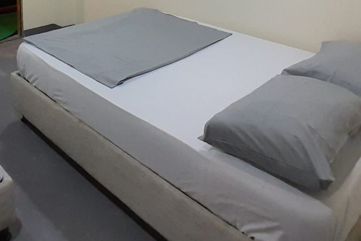 A single bed with a grey mattress and pillows, a white wall, and a door to the left.