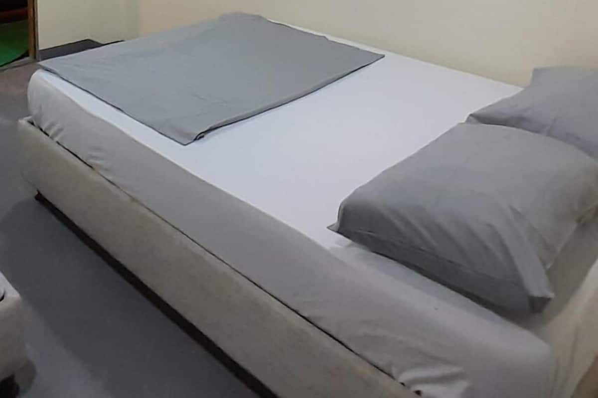 A single bed with a grey mattress and pillows, a white wall, and a door to the left.