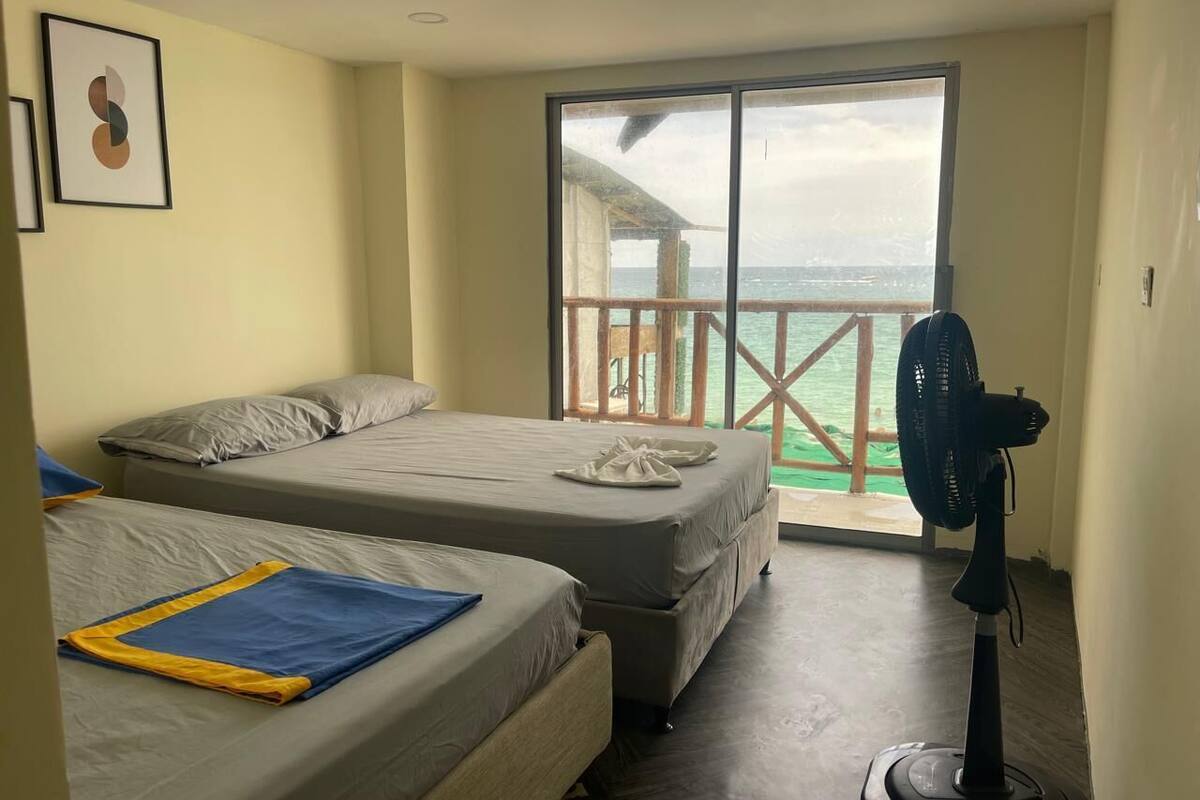 A hotel room with two beds, a balcony with a view of the sea, and a standing fan.