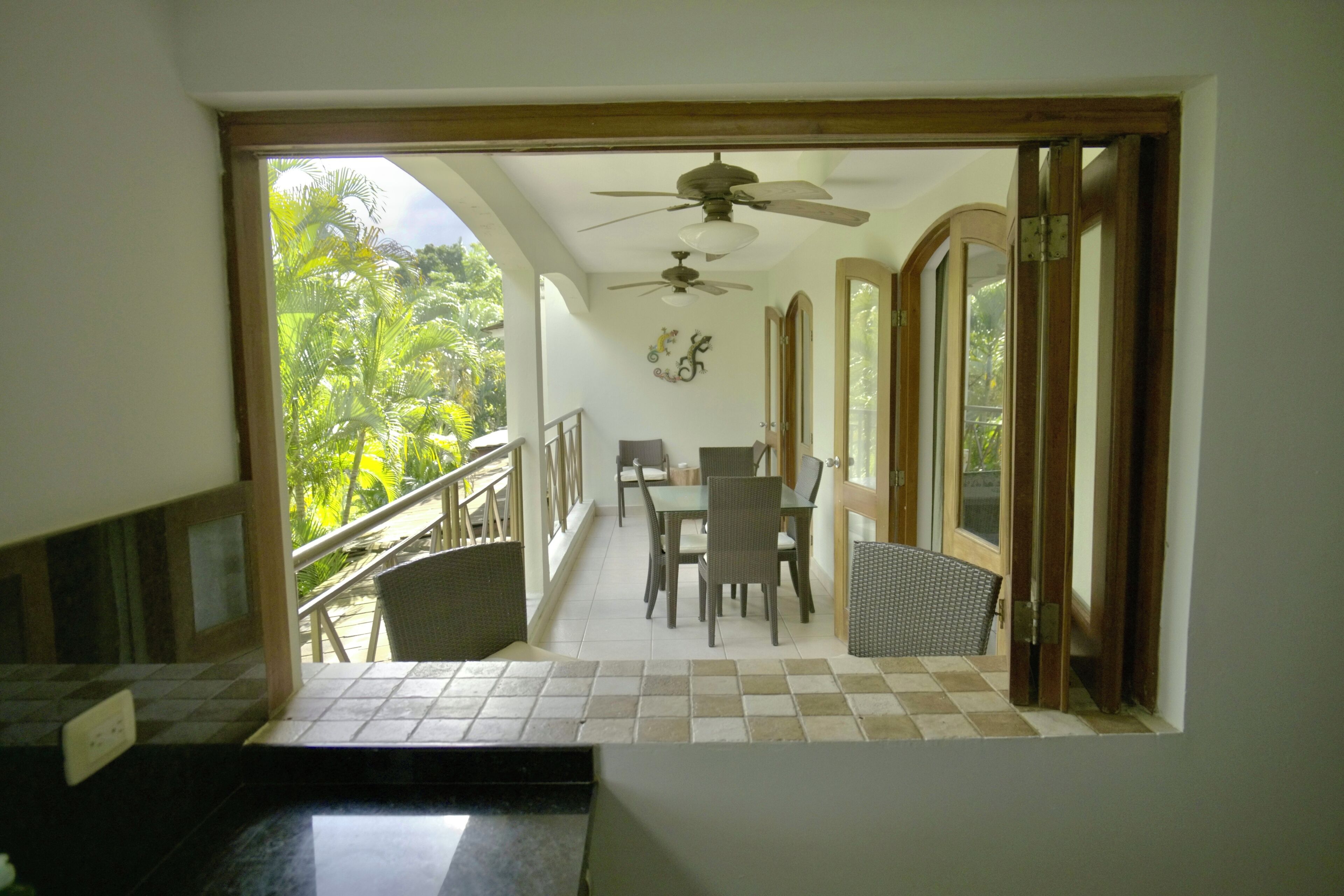 A view from a balcony to a dining area with a ceiling fan, a table, chairs, and a view of greenery outside.