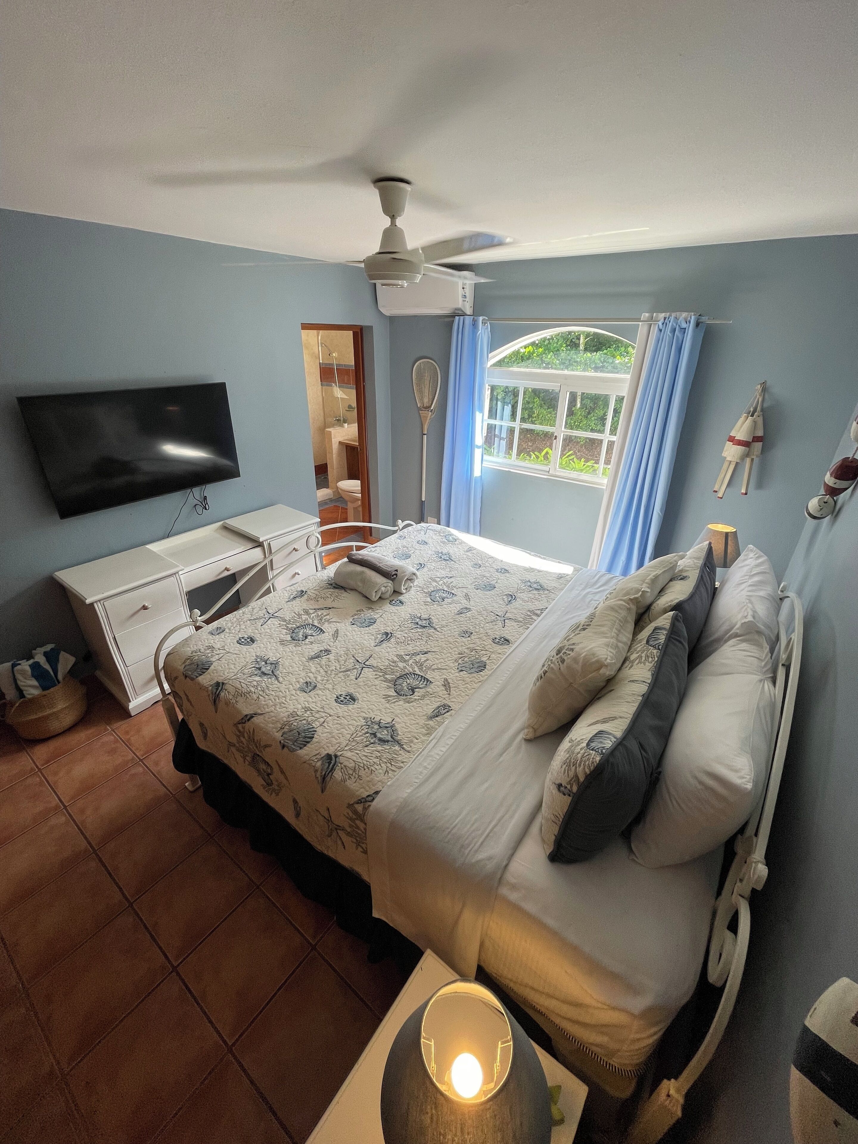 Une chambre à coucher comprenant un lit, un téléviseur, un ventilateur de plafond, une fenêtre avec des rideaux et une lampe de chevet.