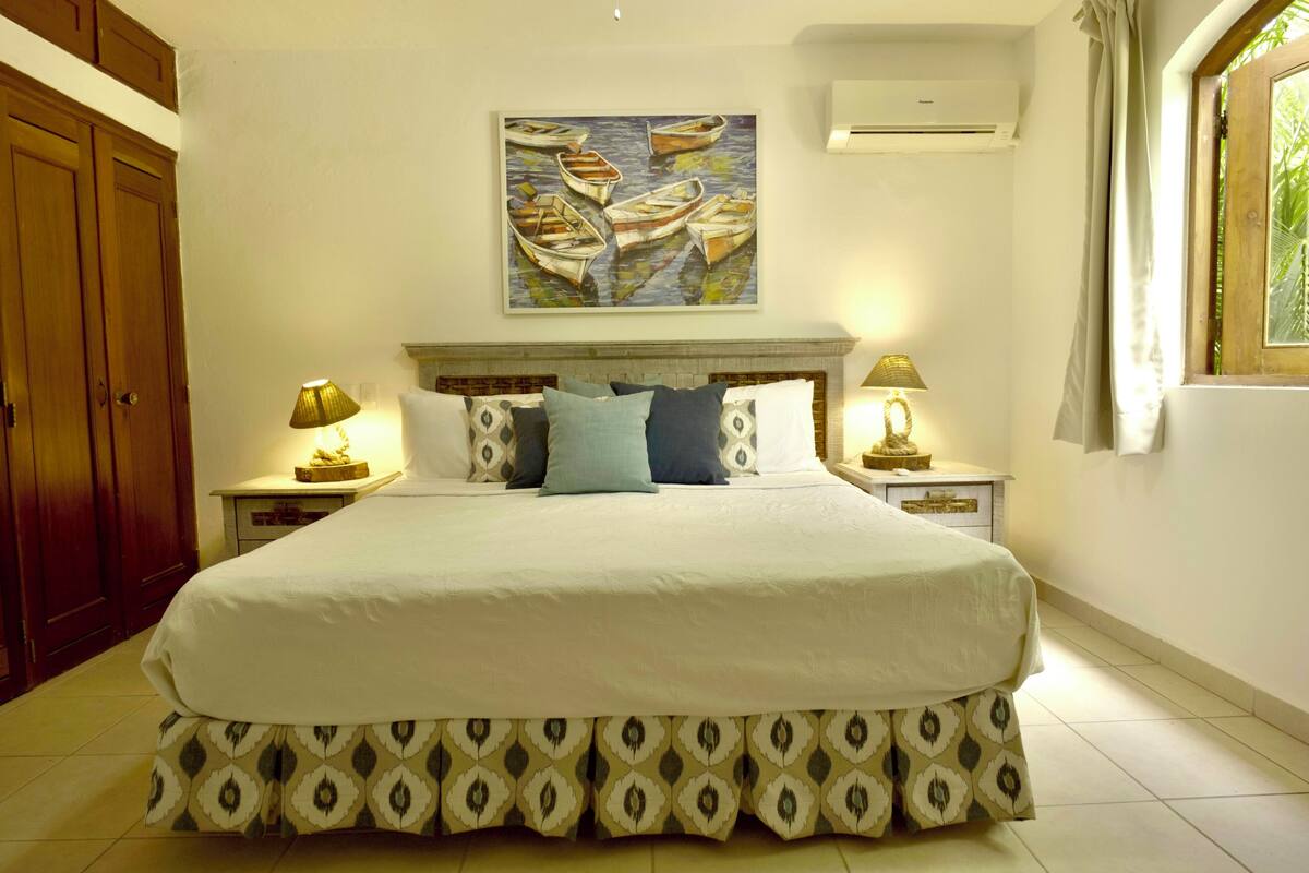 A bedroom with a large bed, two bedside tables with lamps, a wooden wardrobe, a painting of boats, and a window with curtains.