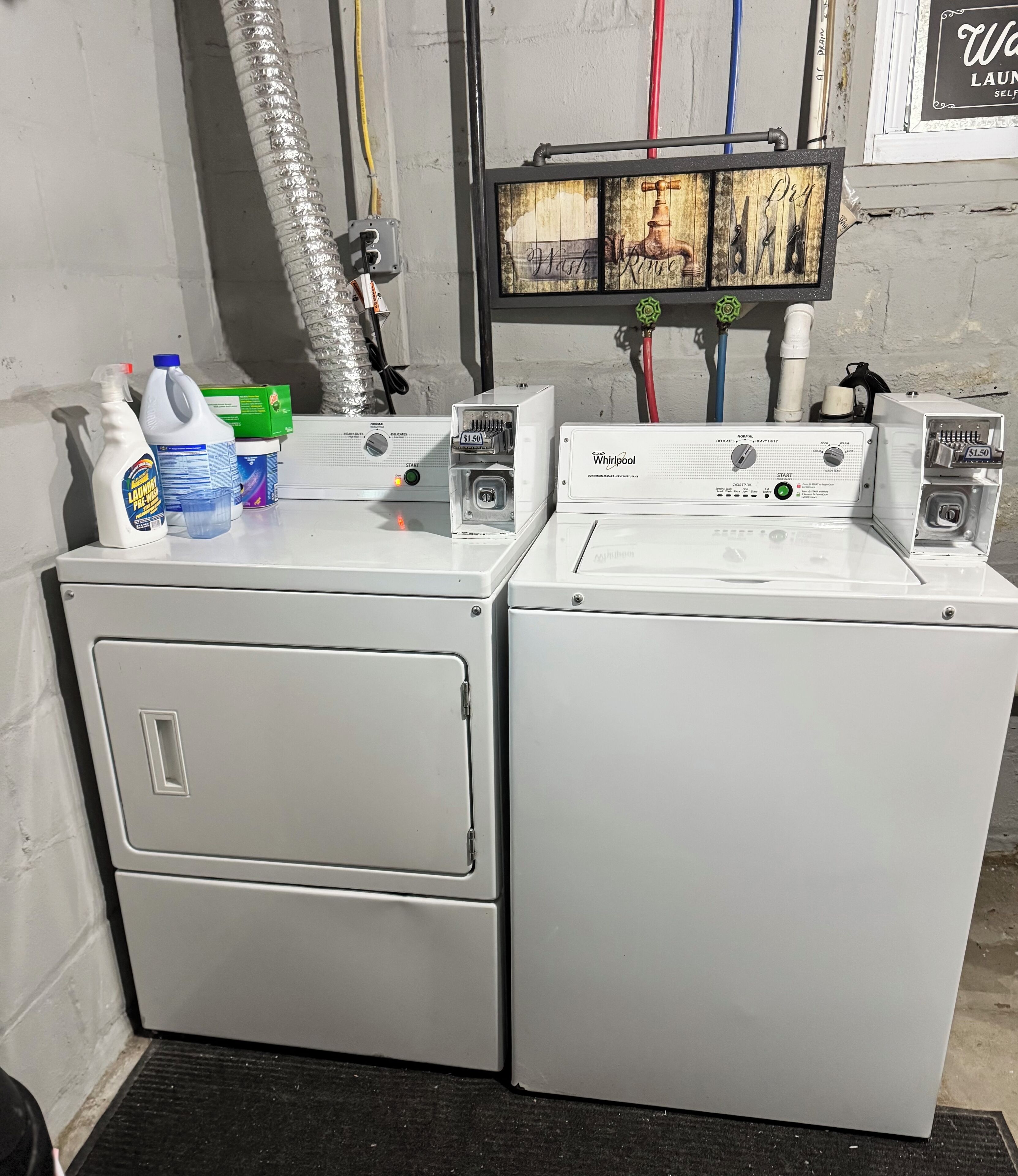 Dos lavadoras Whirlpool de carga frontal con paneles de control, un conducto de ventilación y un letrero en la pared.