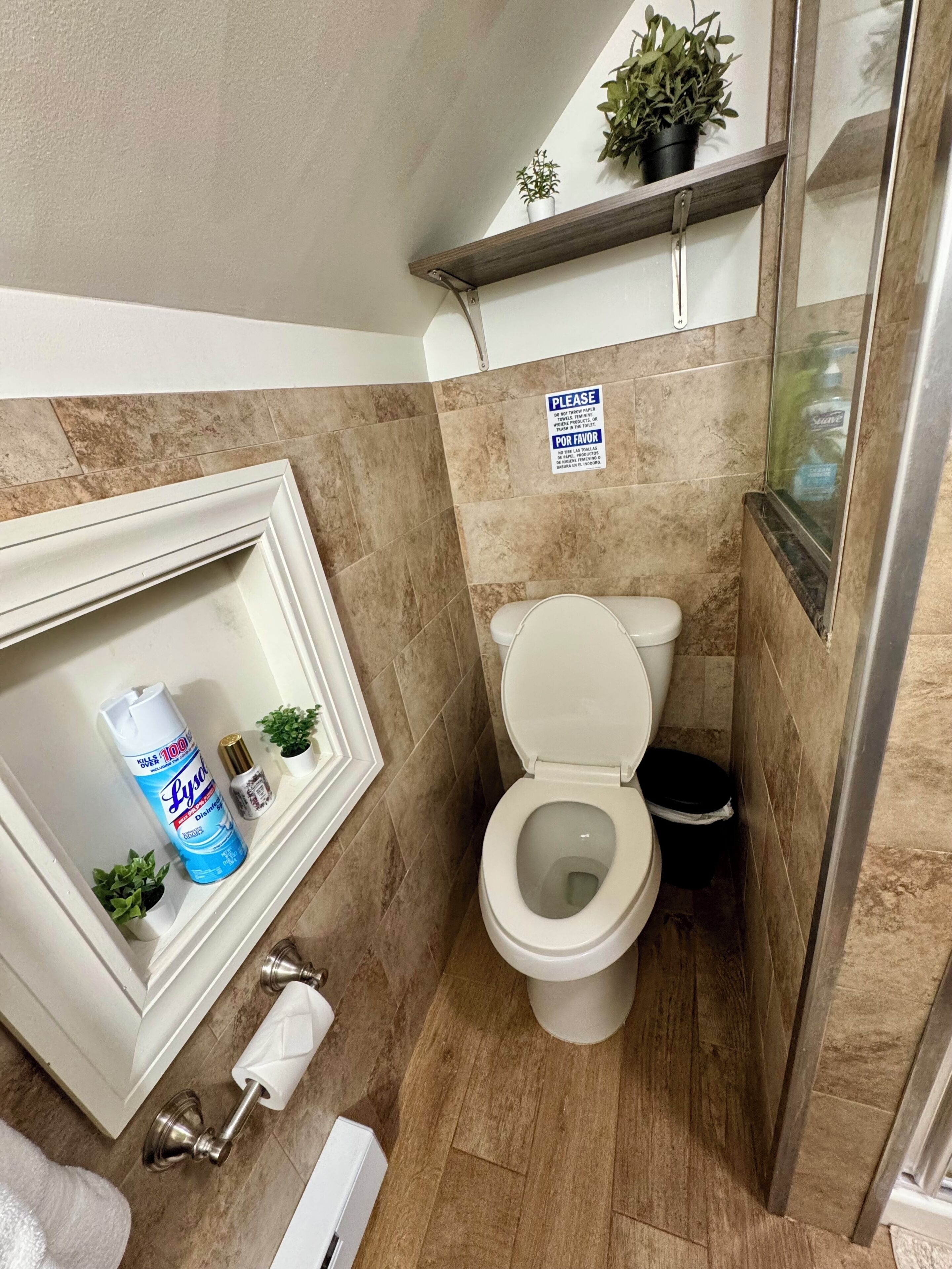 A bathroom with a toilet, a sink with a mirror, a shelf with plants, and a sign on the wall.