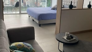 Uma sala de estar moderna com uma cama grande, um sofá, uma mesinha e vista para o horizonte urbano através de amplas janelas.