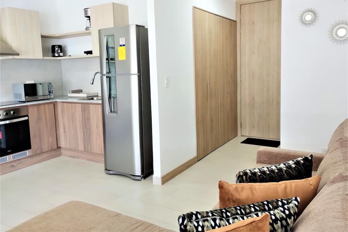 Eine moderne Küche mit einem Edelstahlkühlschrank, Holzschränken und einer Mikrowelle. Im Wohnbereich befindet sich eine Couch mit gemusterten Kissen.