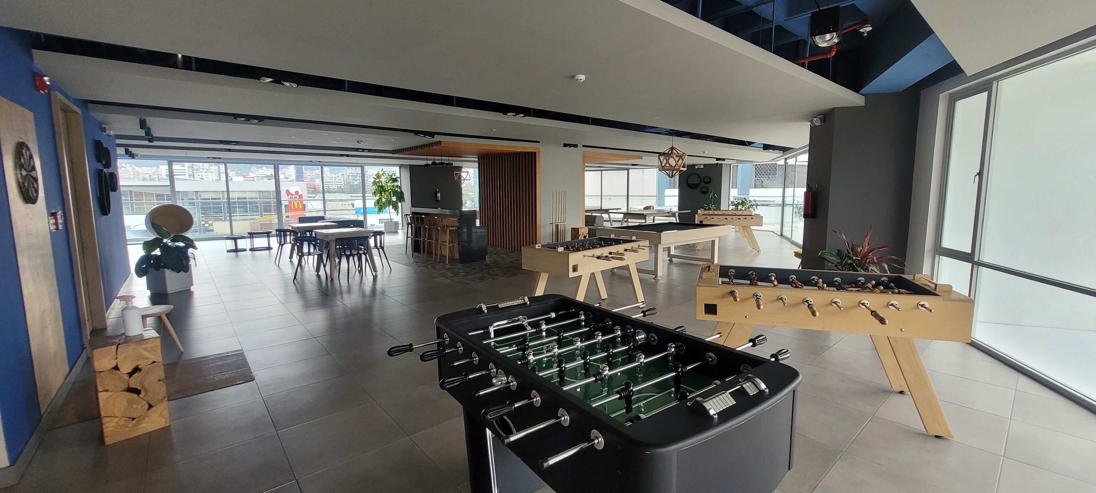 A modern indoor space with two foosball tables, a bar area, and a seating area with tables and chairs.