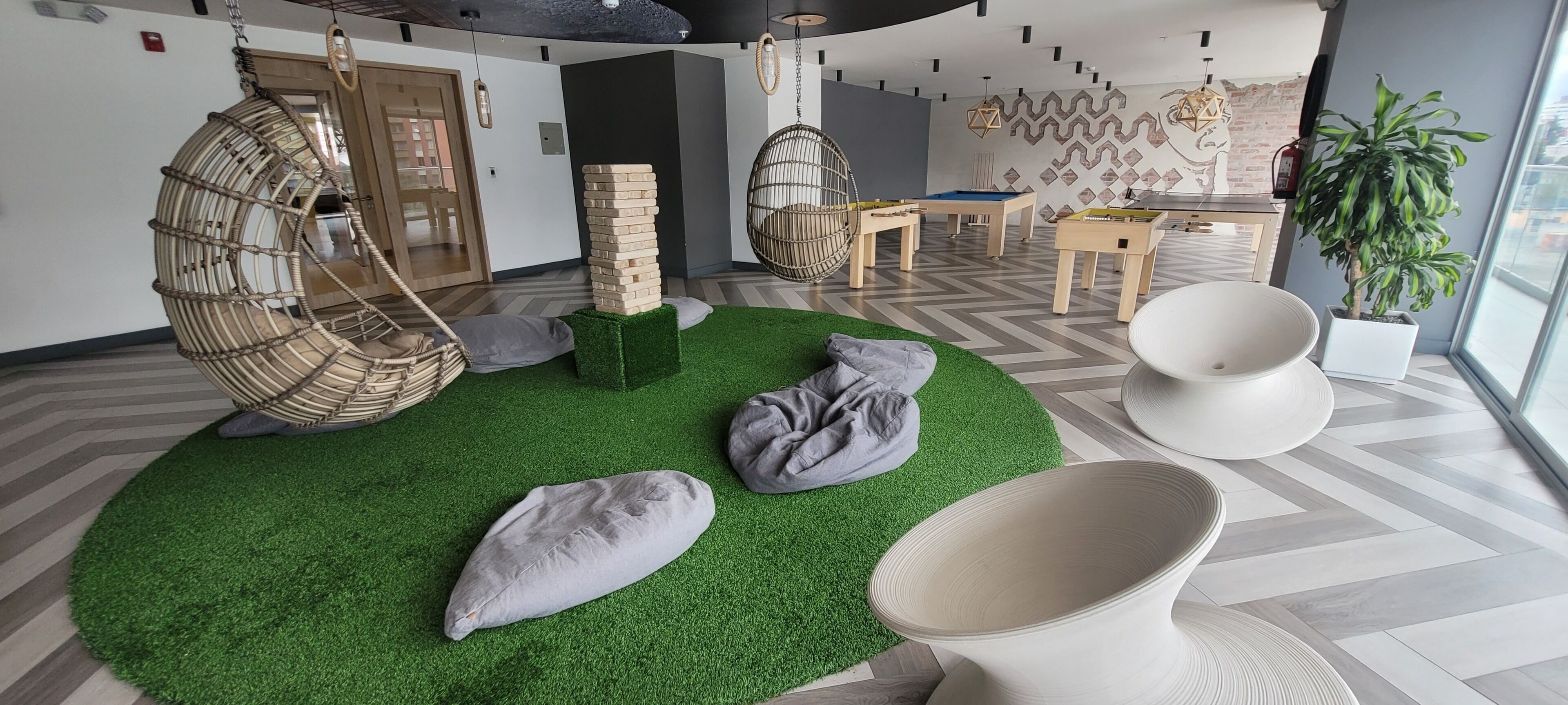 Una habitación moderna con una zona de césped artificial verde, un sillón colgante, una pila de bloques de madera, una silla blanca y una planta en maceta.