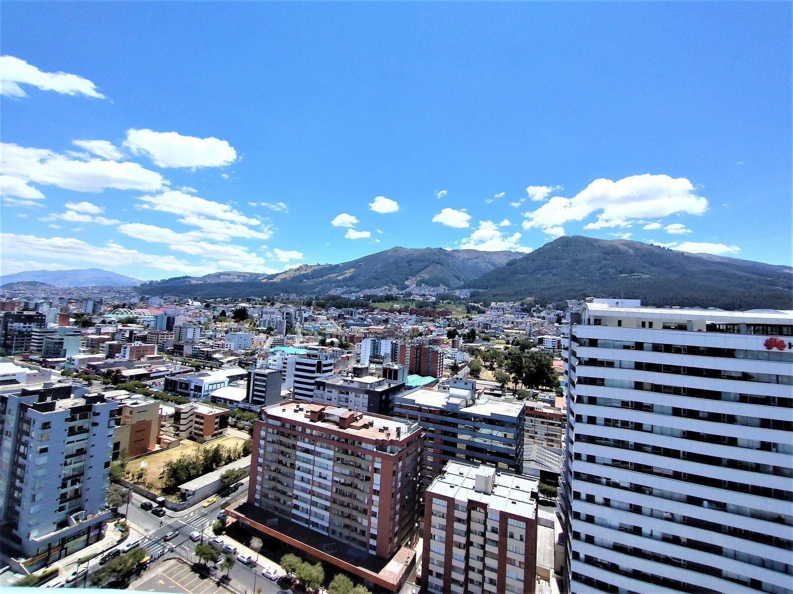 A cityscape with a prominent building featuring a red logo, surrounded by various other buildings and a mountainous backdrop.