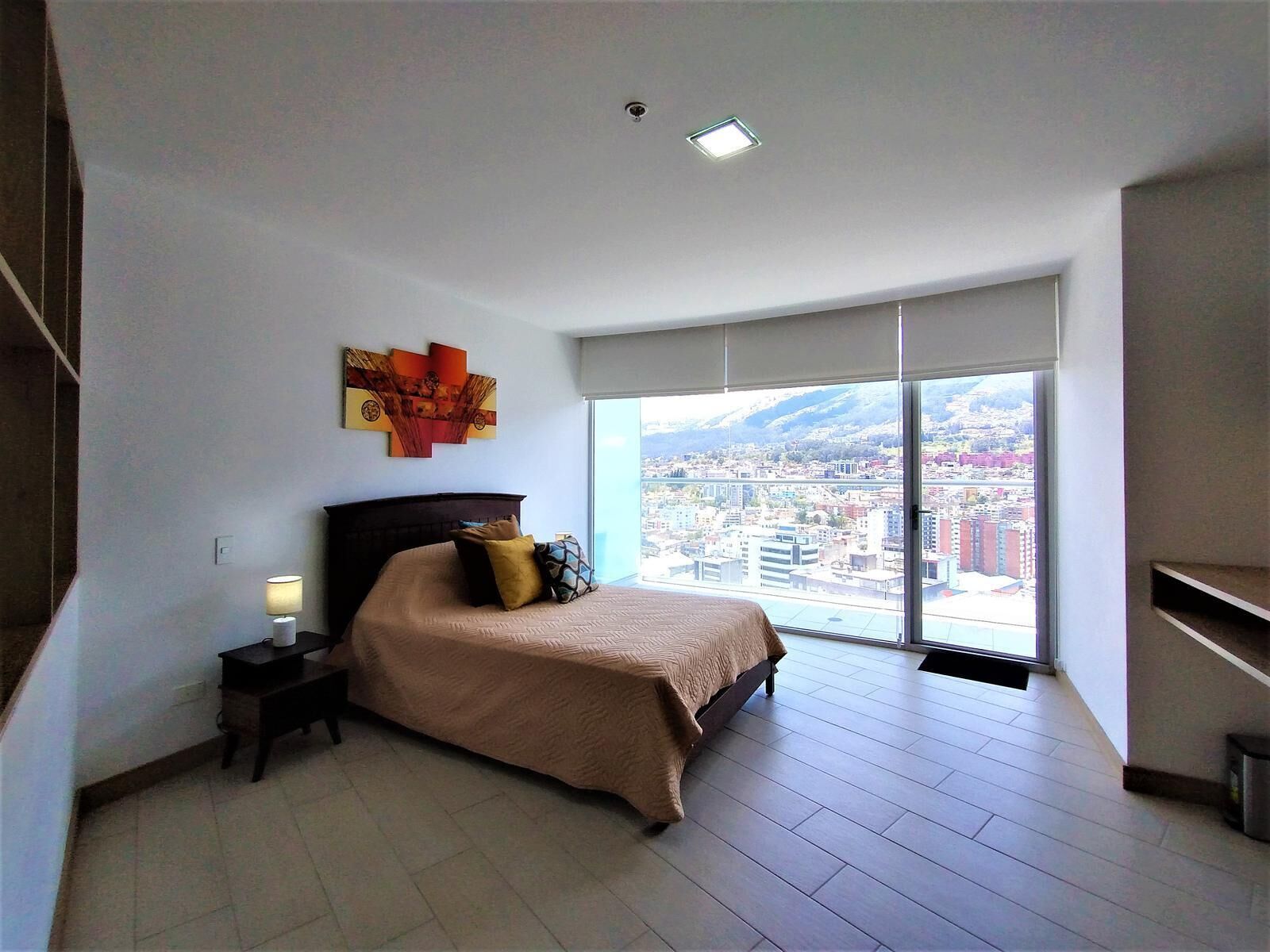 A bedroom with a bed, a nightstand, a lamp, a large window with a city view, and a ceiling light.