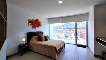 A bedroom with a bed, a nightstand, a lamp, a large window with a city view, and a ceiling light.