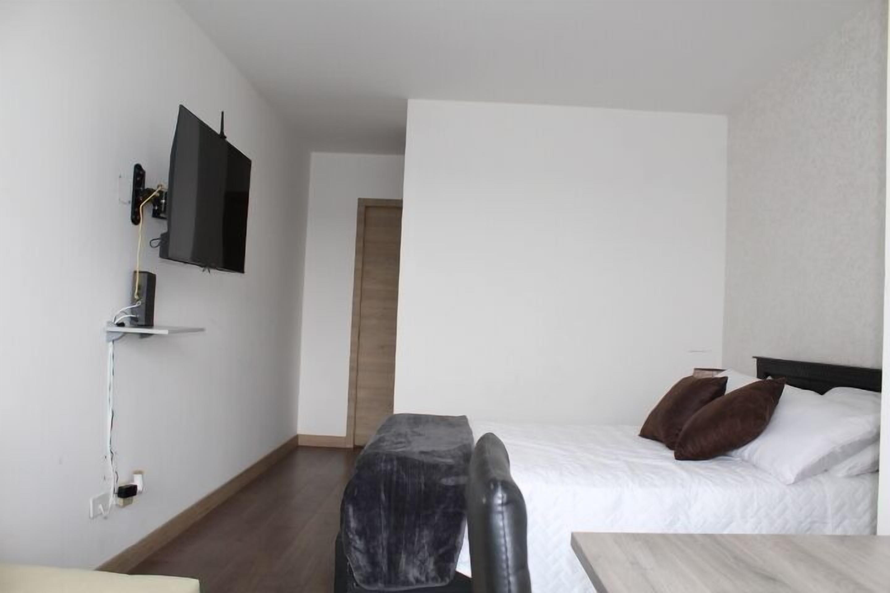 Ett hotellrum med en säng, ett skrivbord, en stol, en tv som är monterad på väggen och en dörr som leder till ett annat rum.