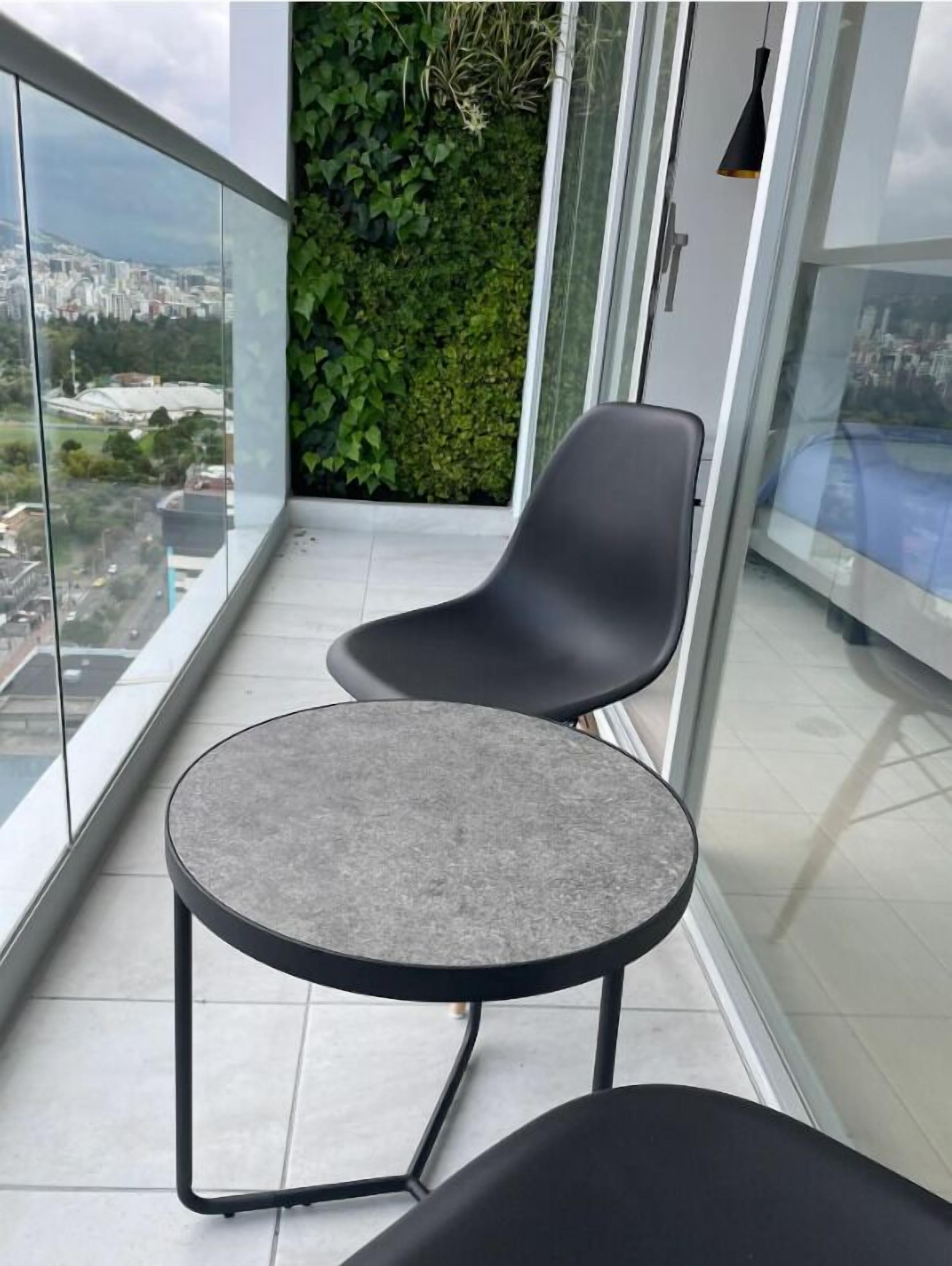 A balcony with a round table and two chairs, a green wall with plants, and a city view through large glass windows.