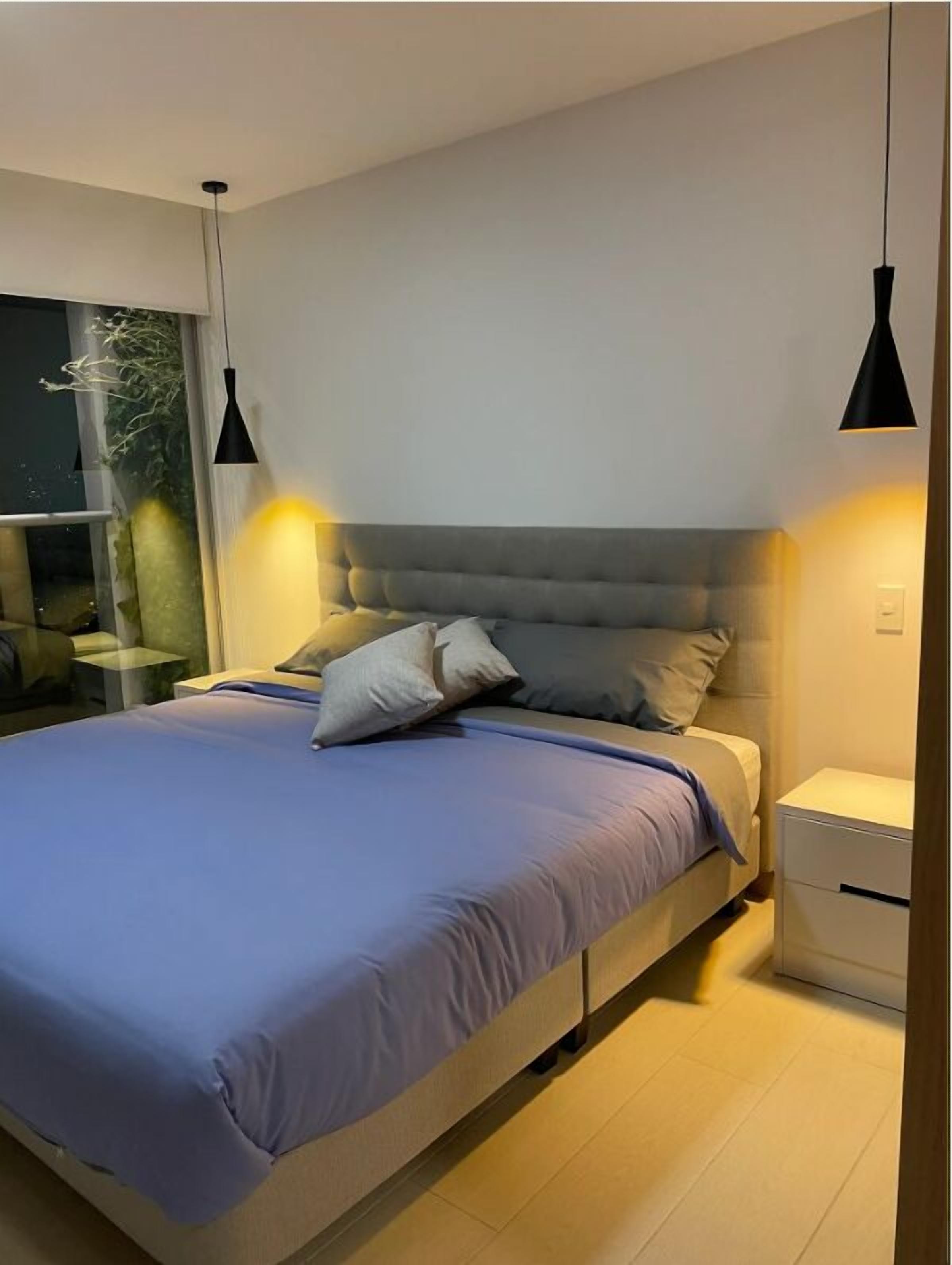 A modern bedroom with a large bed, two bedside lamps, a nightstand, and a window with a view of greenery.