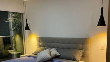 A modern bedroom with a large bed, two bedside lamps, a nightstand, and a window with a view of greenery.