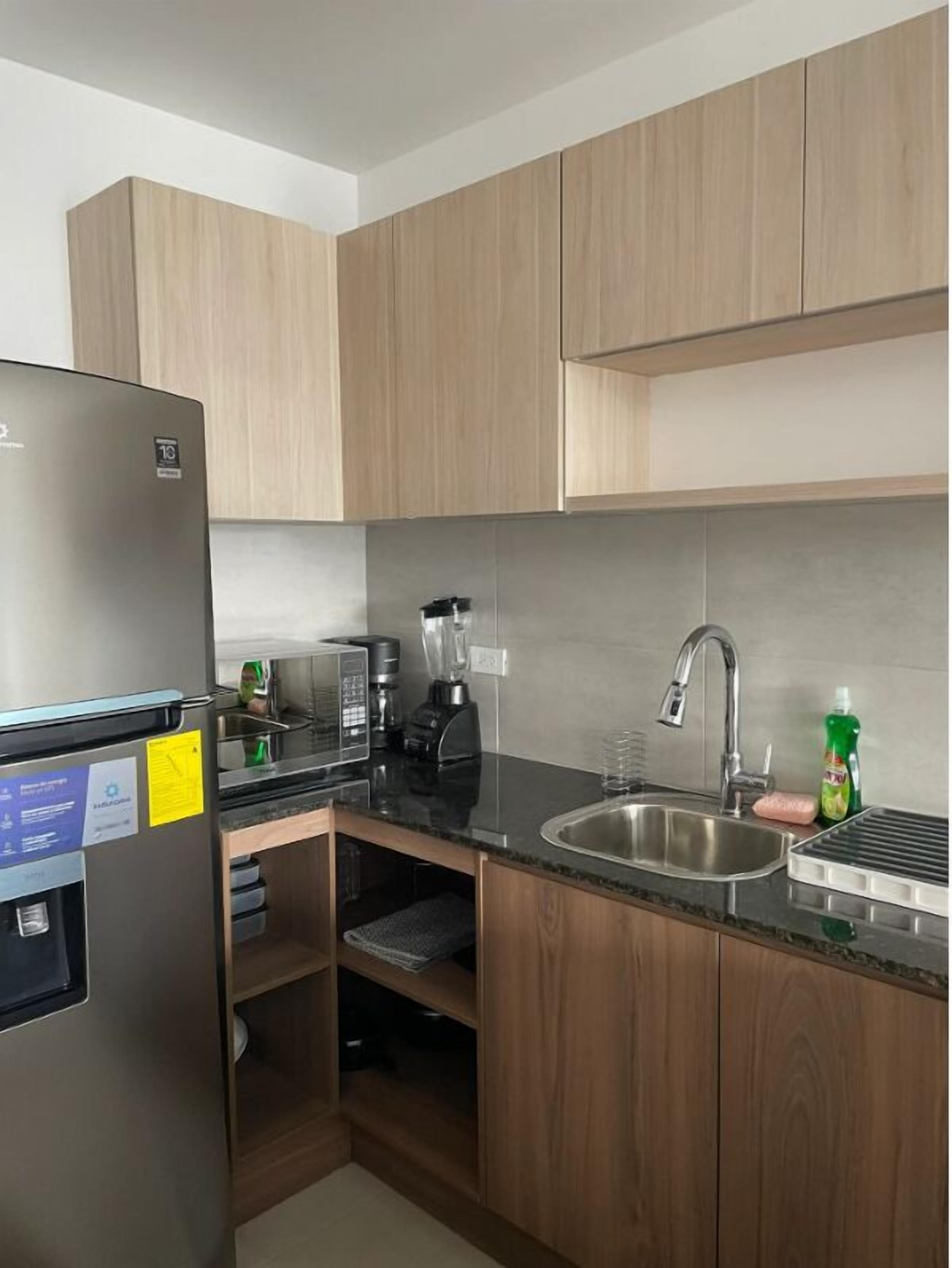 Eine kompakte Küche mit Holzausziehtüren, einem Edelstahlkühlschrank, einer Kaffeemaschine, einem Mixer und einem Spülbecken.