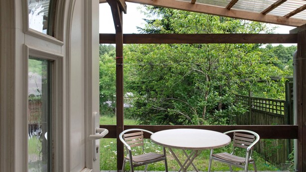Een houten terras met een tafel en twee stoelen, met uitzicht op een tuin met bomen en een houten omheining.