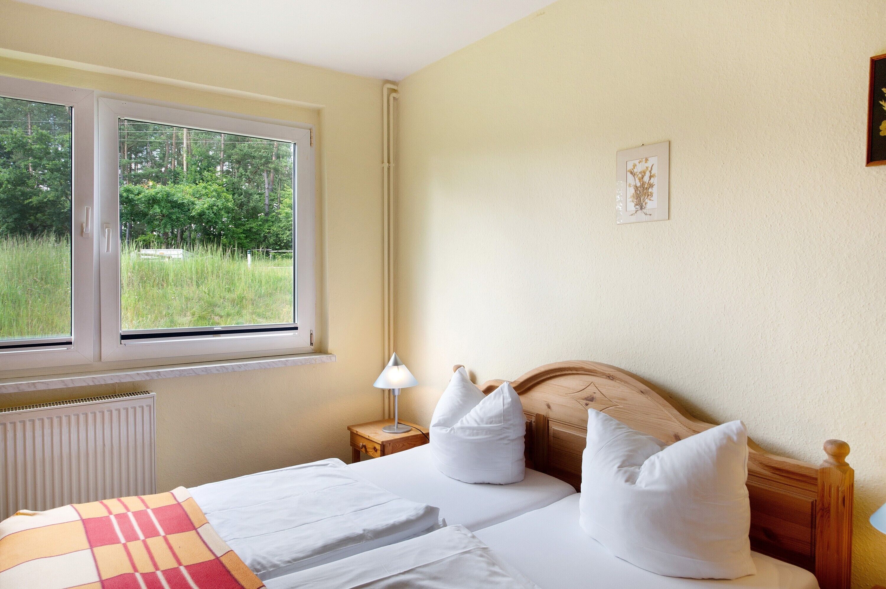 A bedroom with a bed, a nightstand, a lamp, a window with a view of greenery, and a framed picture on the wall.