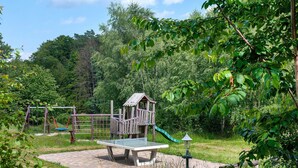 Un parque infantil con estructura de juego de madera, columpios y un tobogán, rodeado de follaje.