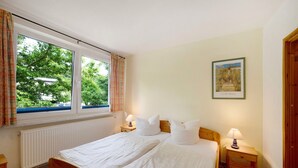 Une chambre à coucher avec un lit, deux tables de chevet, une fenêtre avec des rideaux, un radiateur et un tableau encadré au mur.