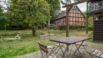A rustic outdoor seating area with a wooden table and benches, a picnic table, and a traditional house in the background.