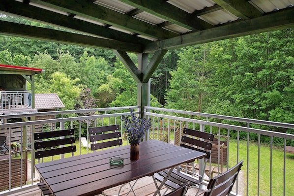 Une terrasse extérieure couverte, dotée d’une table et de chaises, d’une rambarde et offrant une vue sur une zone boisée.