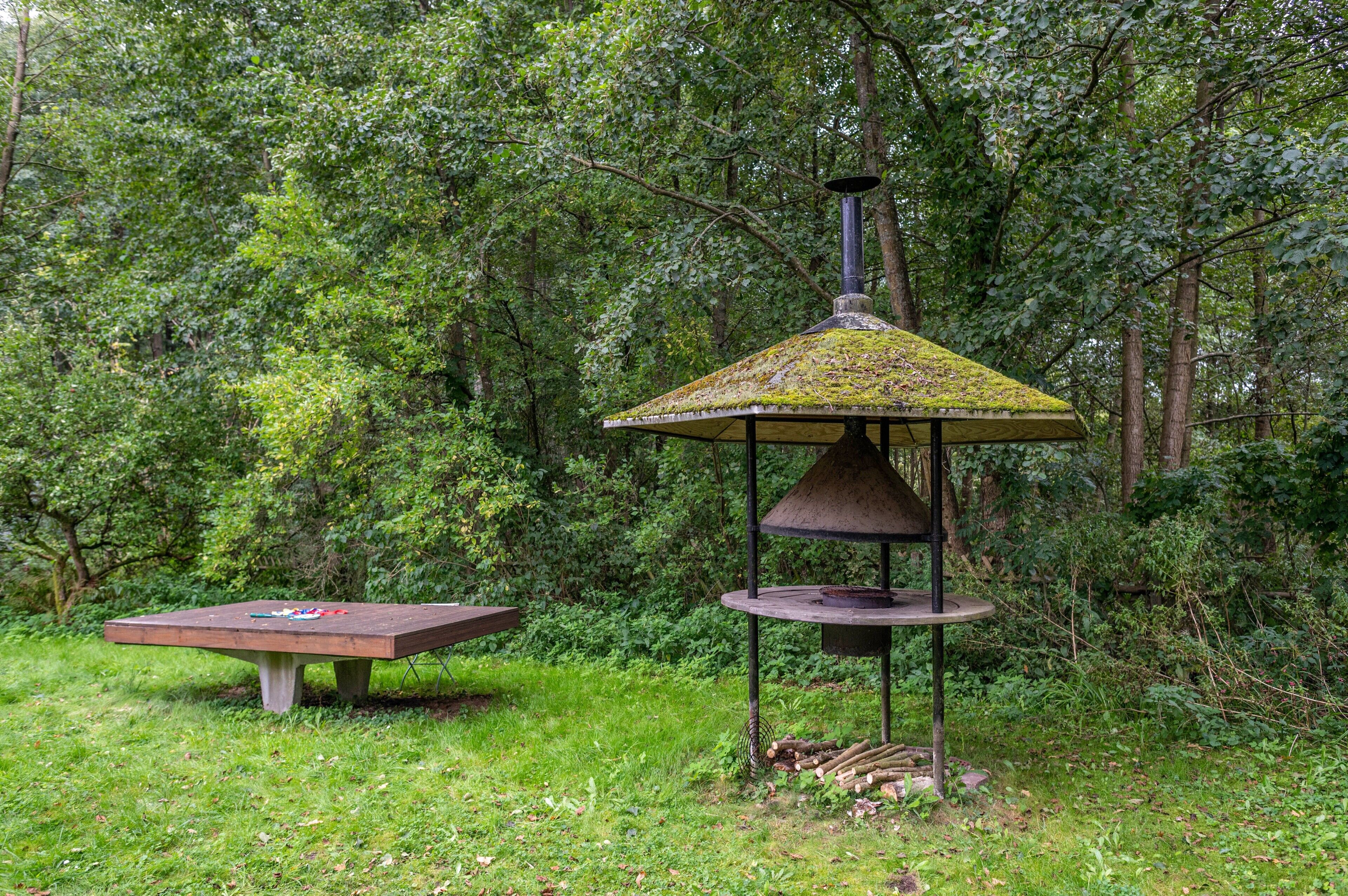 Een prieel met een kegelvormig dak, een picknicktafel en een bebost achtergrond.