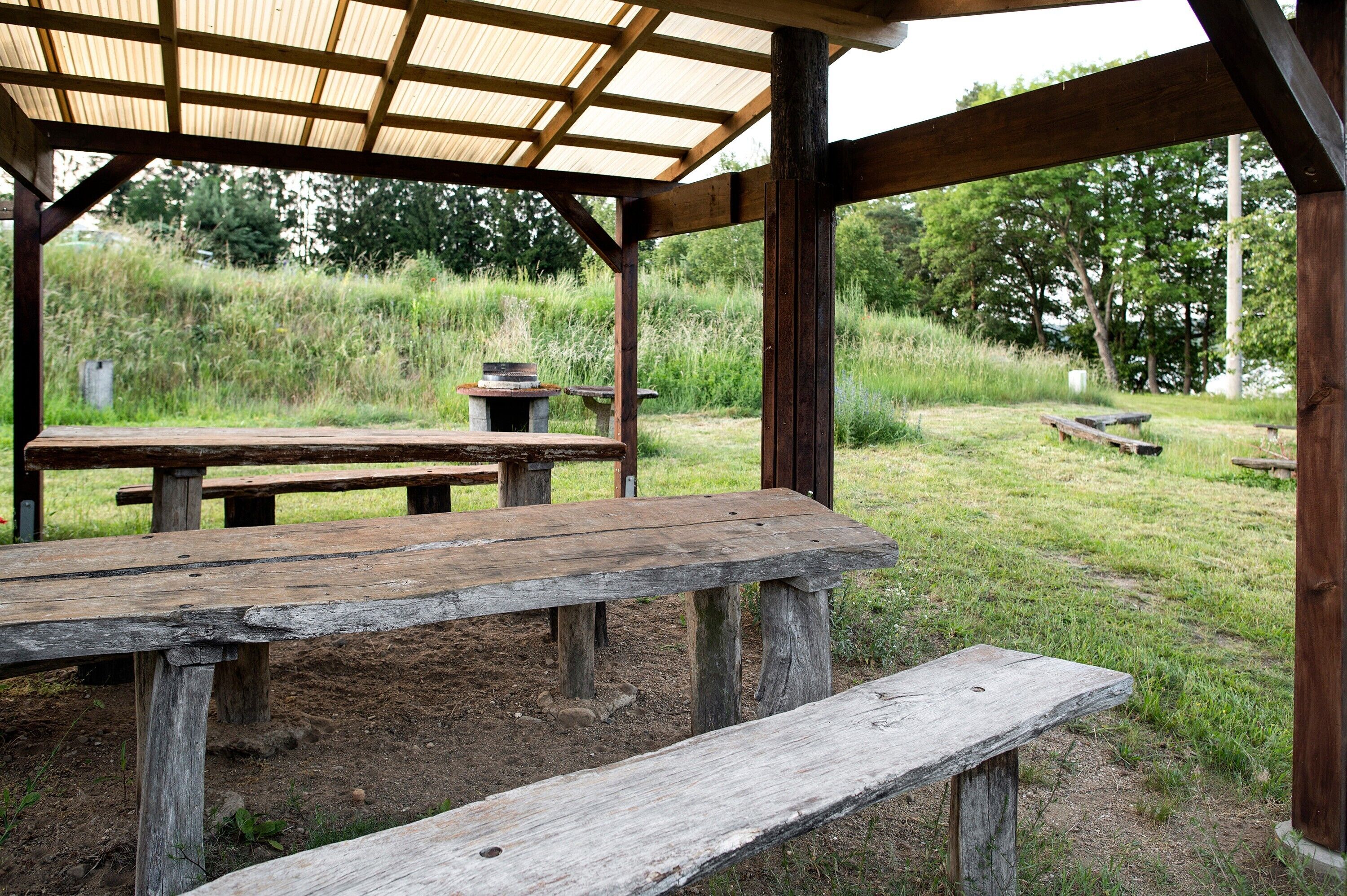 A rustic wooden pavilion with benches and a table, surrounded by a grassy field and trees.