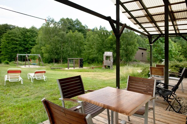 Eine Terrasse im Freien mit Holztisch und -stühlen, einer Schaukel und einem kleinen Geräteschuppen auf einer Rasenfläche, im Hintergrund Bäume.