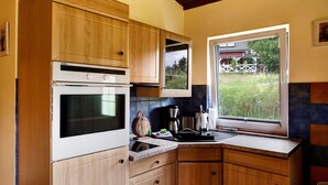 A kitchen with wooden cabinets, a built-in oven, a window with a view of a barn, and a countertop with various appliances. - Holiday home 'Rethra Nord' with private terrace and WLAN (Feldberger Seenlandschaft)