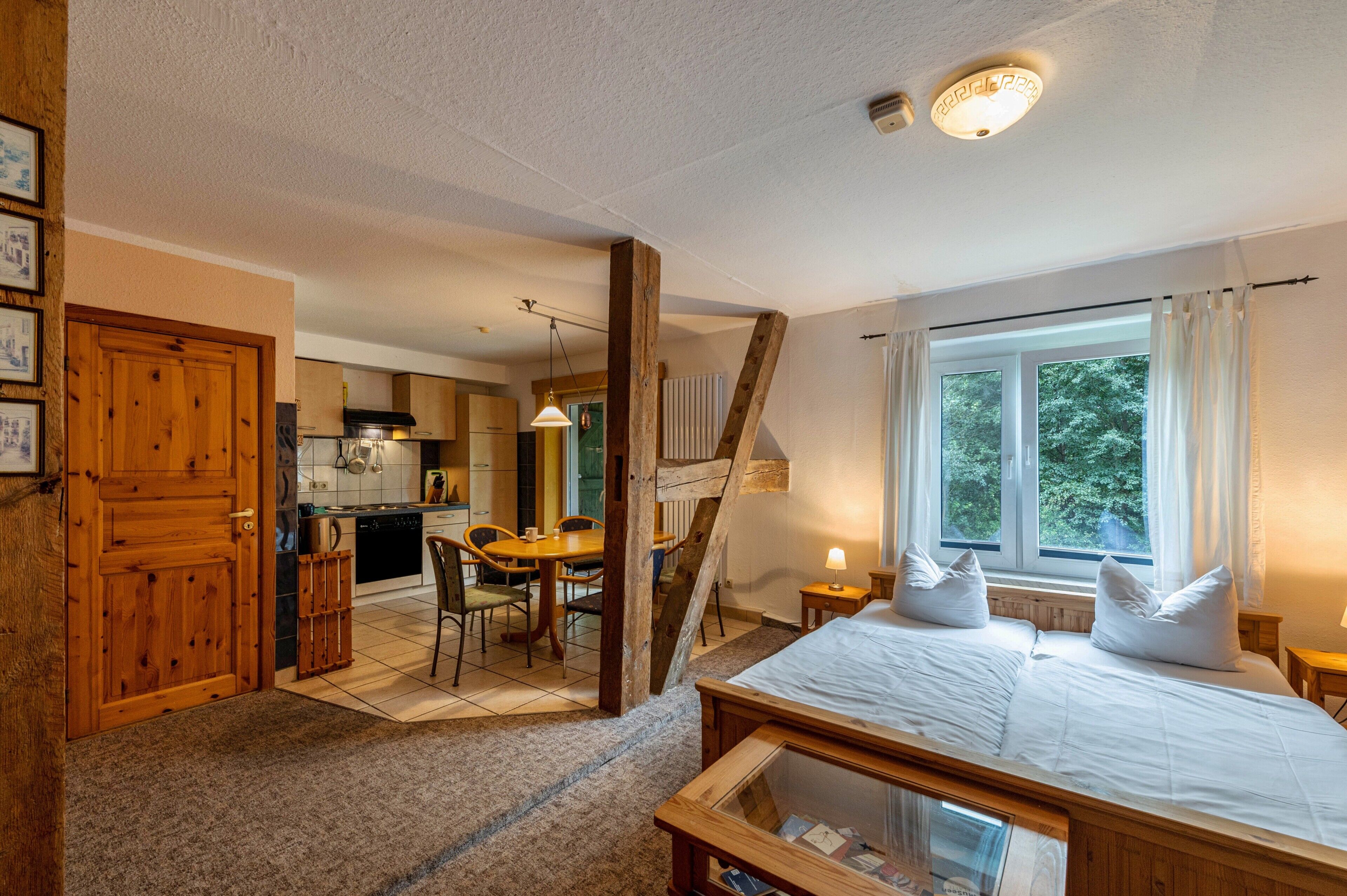 A room with a bed, a window with curtains, a wooden door, and a kitchen area with a dining table.