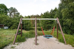 A wooden swing set with a blue swing seat, surrounded by greenery and a sandy path.